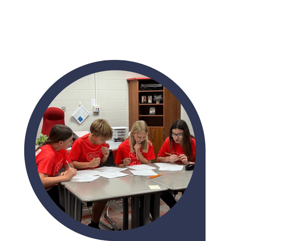 Four students in red shirts work on worksheets around a classroom table, concentrating on their papers.